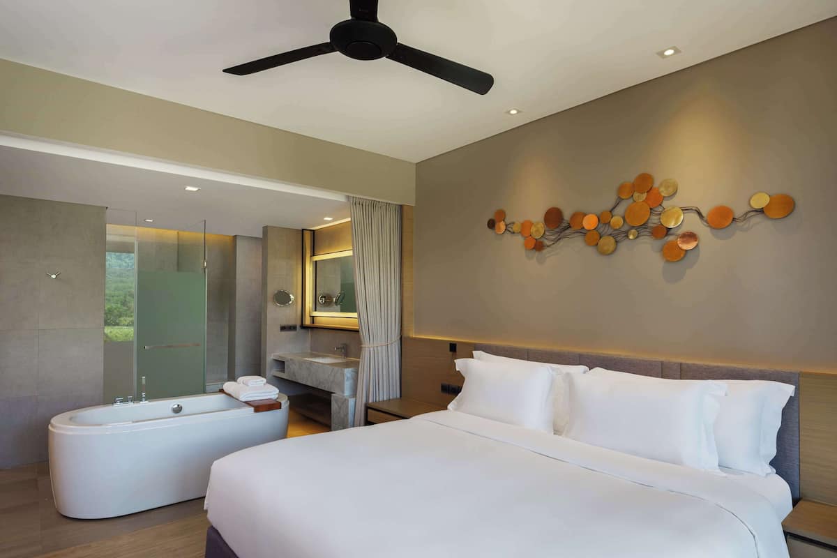 A modern hotel room with a large bed, a separate bathroom with a bathtub, a ceiling fan, and a decorative wall piece.