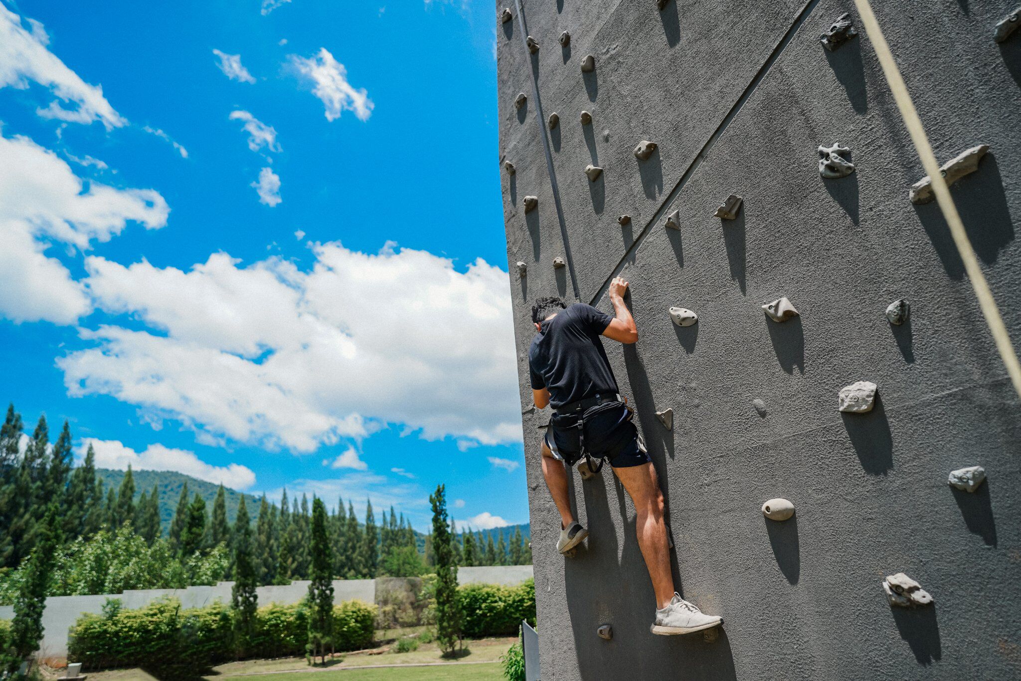 A person climbing a rock wall with handholds and footholds, surrounded by greenery and a clear sky.