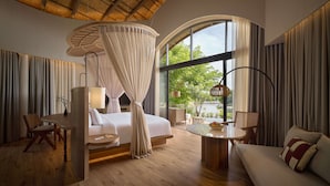 A modern hotel room with a large bed, a sitting area with a sofa and a table, a dining table, and a large window offering a view of greenery outside. - dusitD2 Khao Yai (Pak Chong)