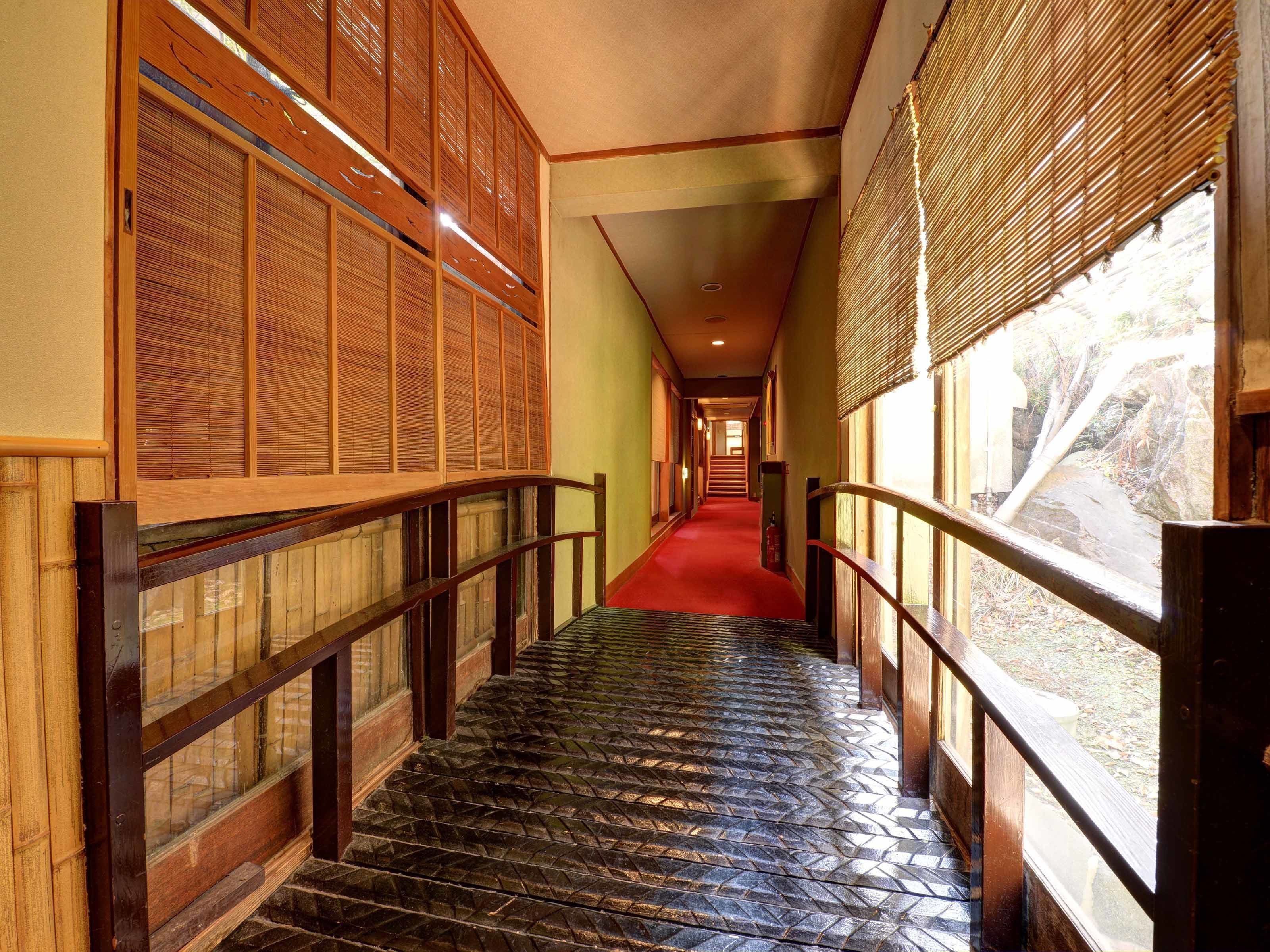 A corridor with wooden railings, a red carpet, and large windows with bamboo blinds.