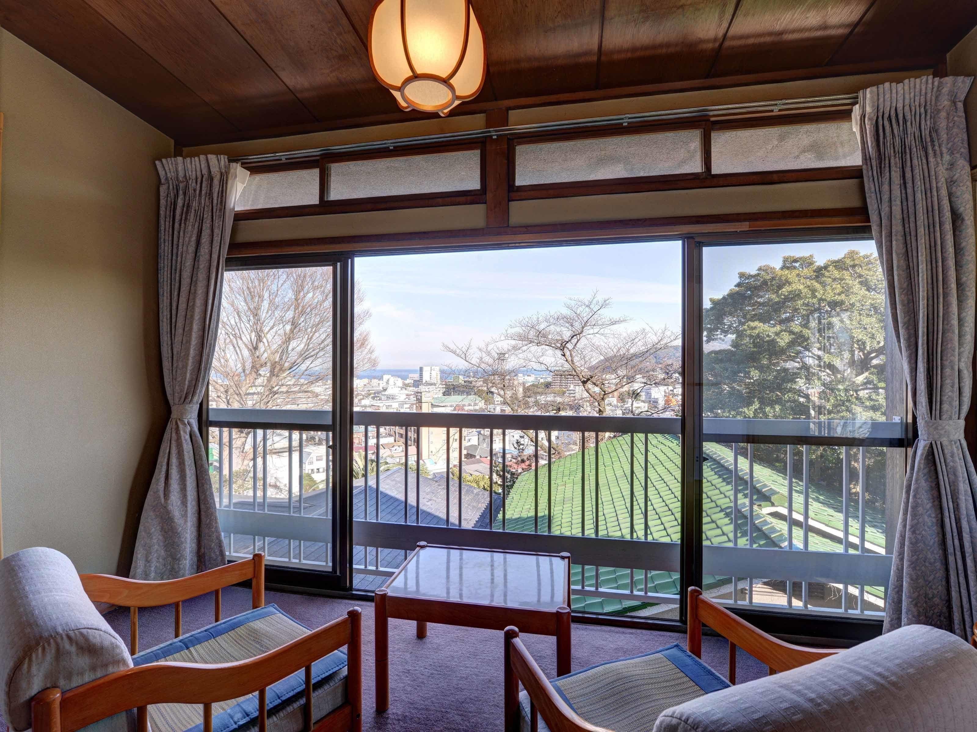 A room with a large window, wooden furniture, and a view of a cityscape and greenery.
