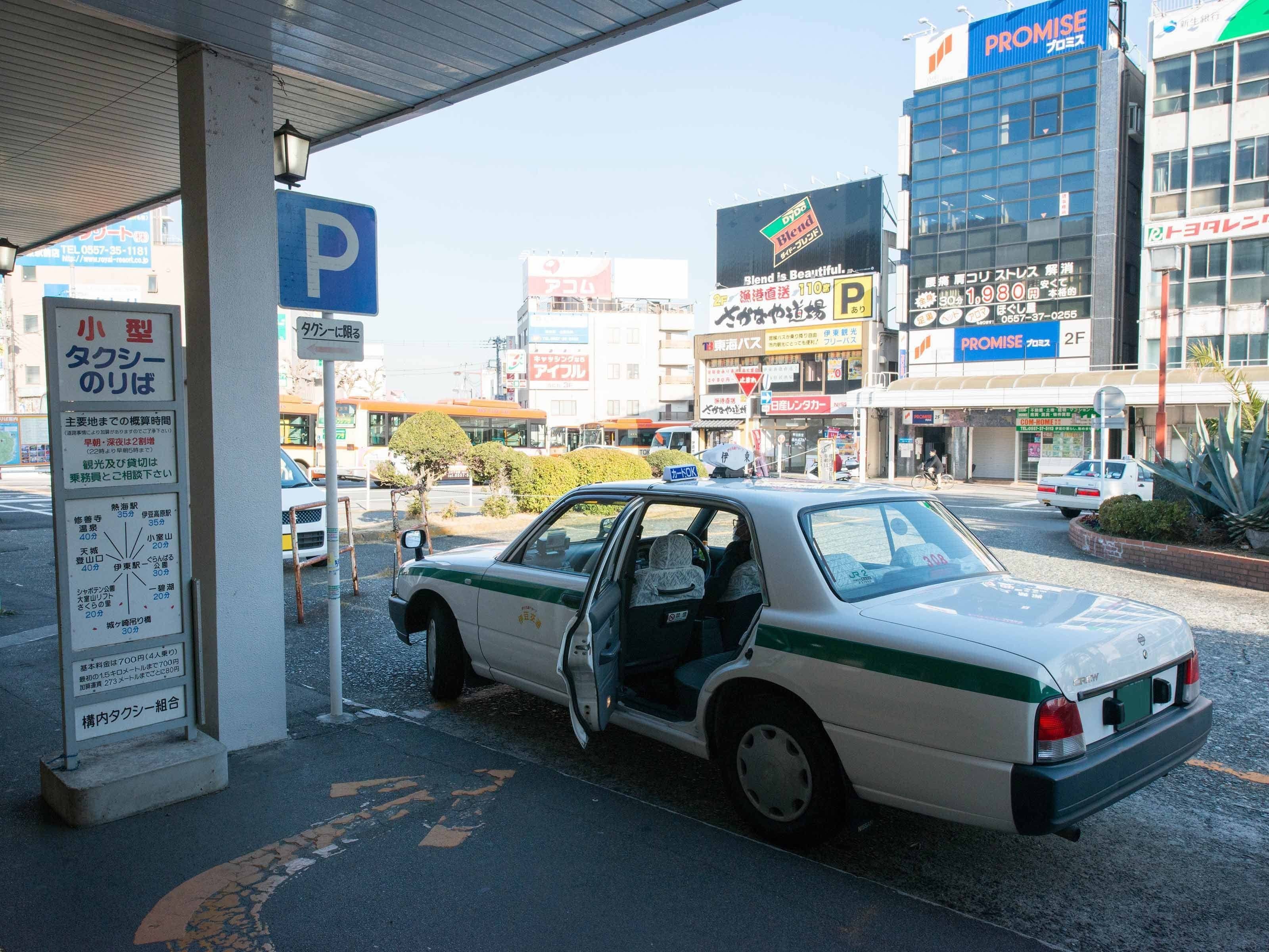 Een witte auto geparkeerd bij een tankstation, met een blauw parkeersbord en een grote reclame voor 'PROMISE' op de achtergrond.