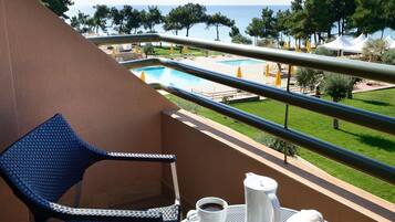A balcony with a table set for two, a chair, and a view of a pool and greenery.