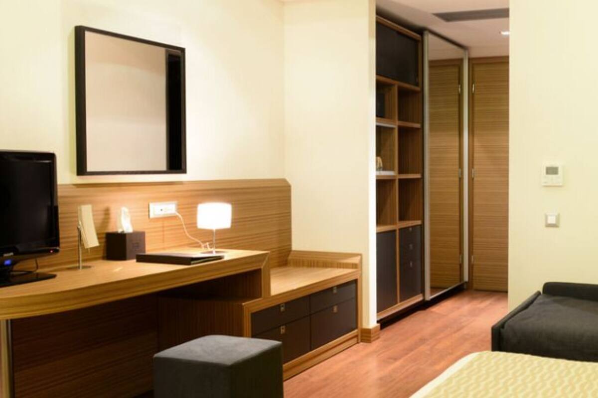 A hotel room with a bed, a desk with a computer, a TV, a bedside table, a lamp, a wardrobe, and a sofa.
