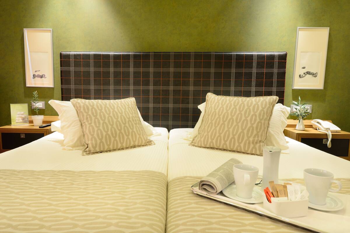 A hotel room with two beds, a large headboard, bedside tables, a mirror, and a tray with a newspaper, cups, and a phone.