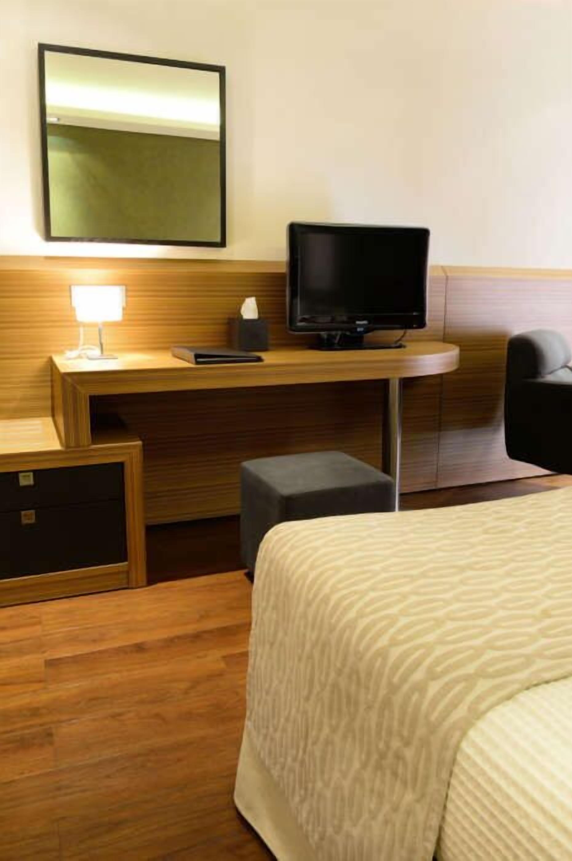 A hotel room with a bed, a desk with a computer and lamp, a chair, and a small cabinet.