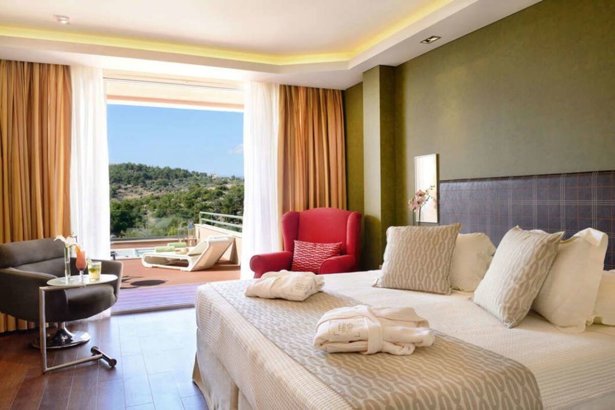 A hotel room with a bed, a red armchair, a black armchair, a small table with a drink, and a view of the outdoors.