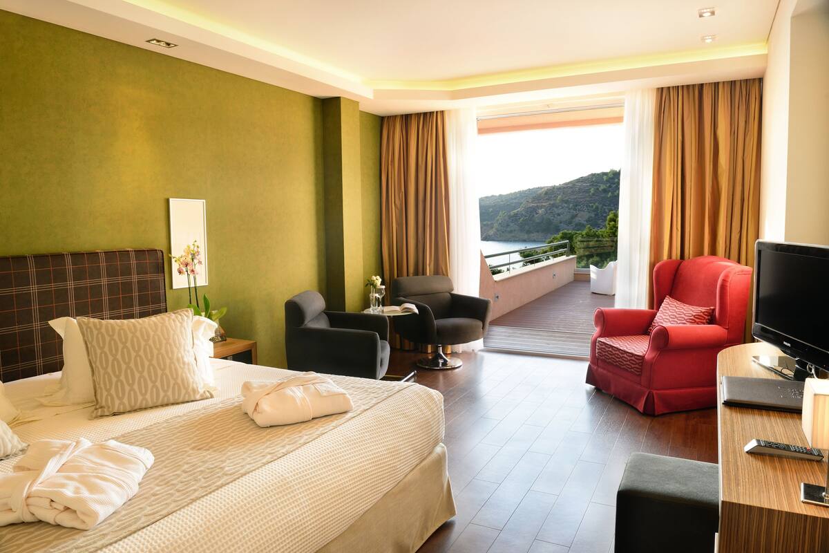 A hotel room with a bed, a red armchair, a television, and a balcony with a view of the hills.
