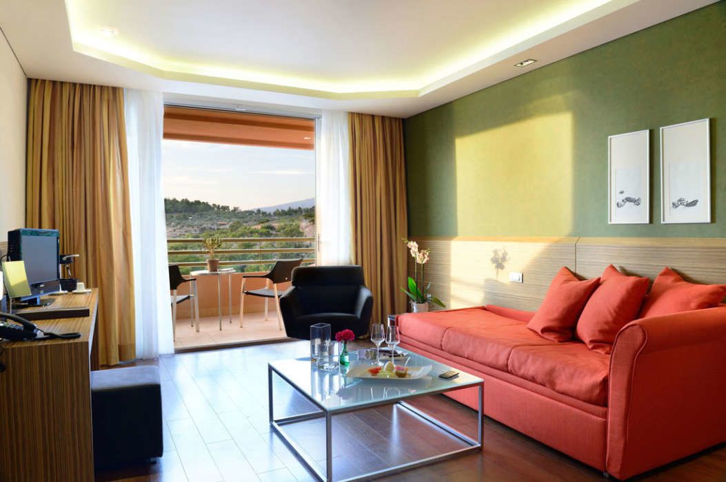 A modern hotel room with a red sofa, a glass coffee table, a desk with a computer, and a balcony with a view of the countryside.