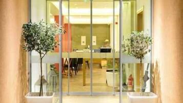 Entrada de hotel con puerta de cristal, plantas en macetas y suelo de baldosas.