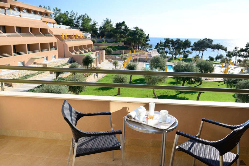 A balcony with two wicker chairs and a small table with a tray of breakfast items. In the background, there is a pool area with yellow umbrellas and a view of the sea and a building with multiple balconies.