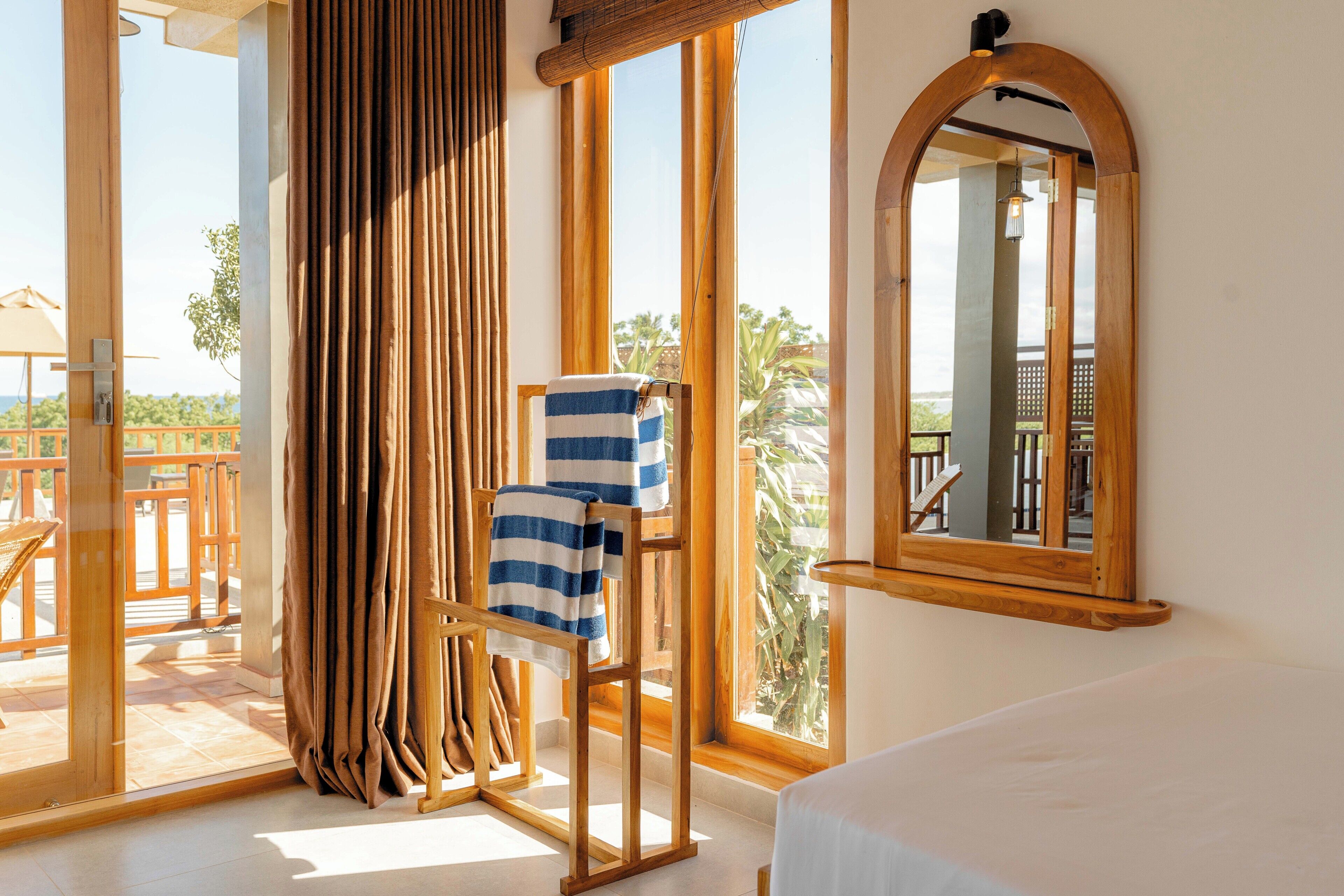 A room with a bed, a wooden chair with a towel, a window with curtains, and a mirror on the wall.