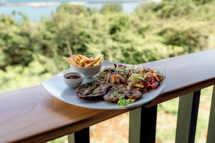 Un plato de comida con papas fritas, ensalada y carne a la parrilla sobre una terraza de madera, con vista a árboles y agua.