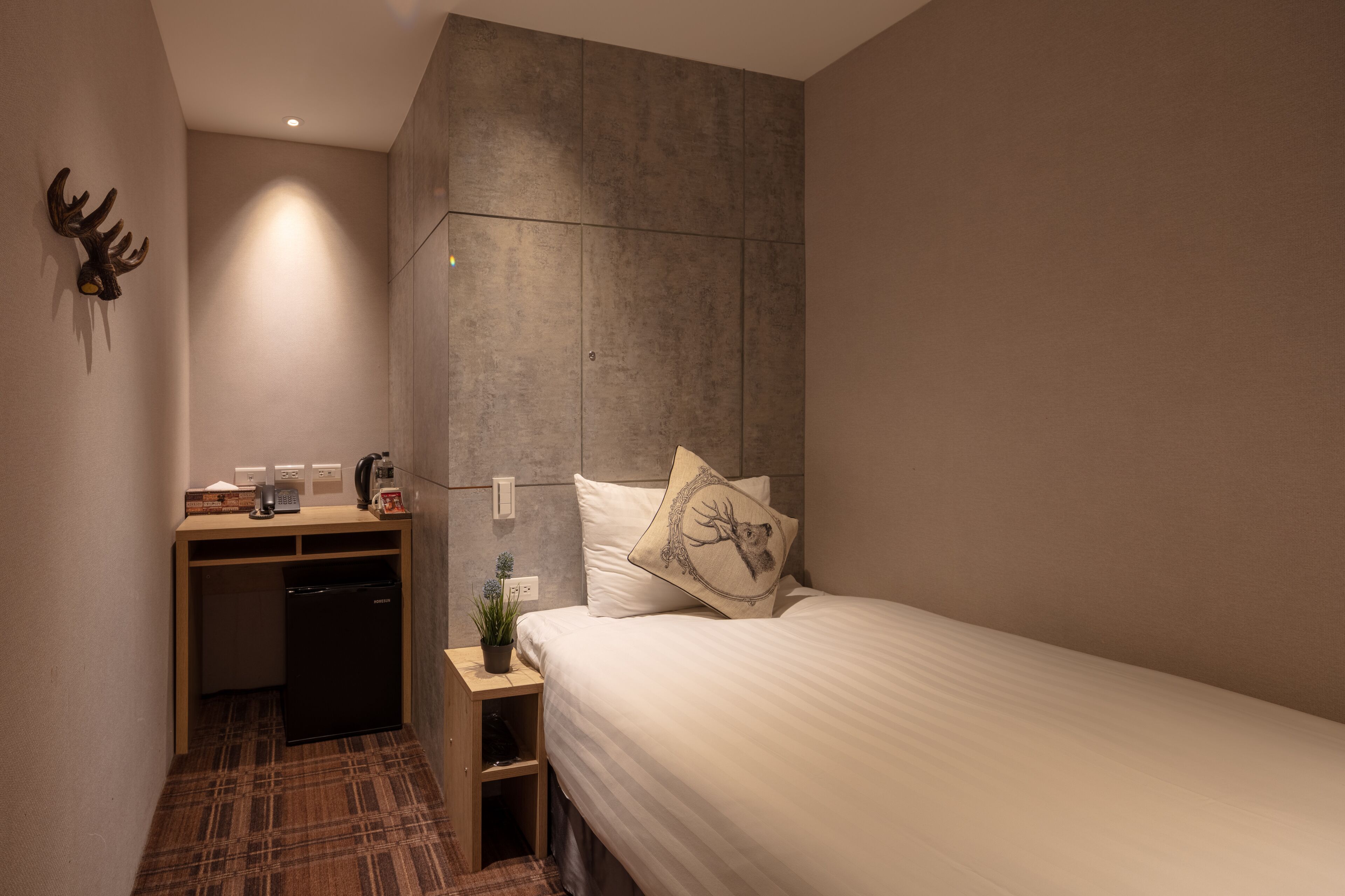 A compact hotel room with a bed, a desk with a computer, a small refrigerator, and a wall-mounted plant.