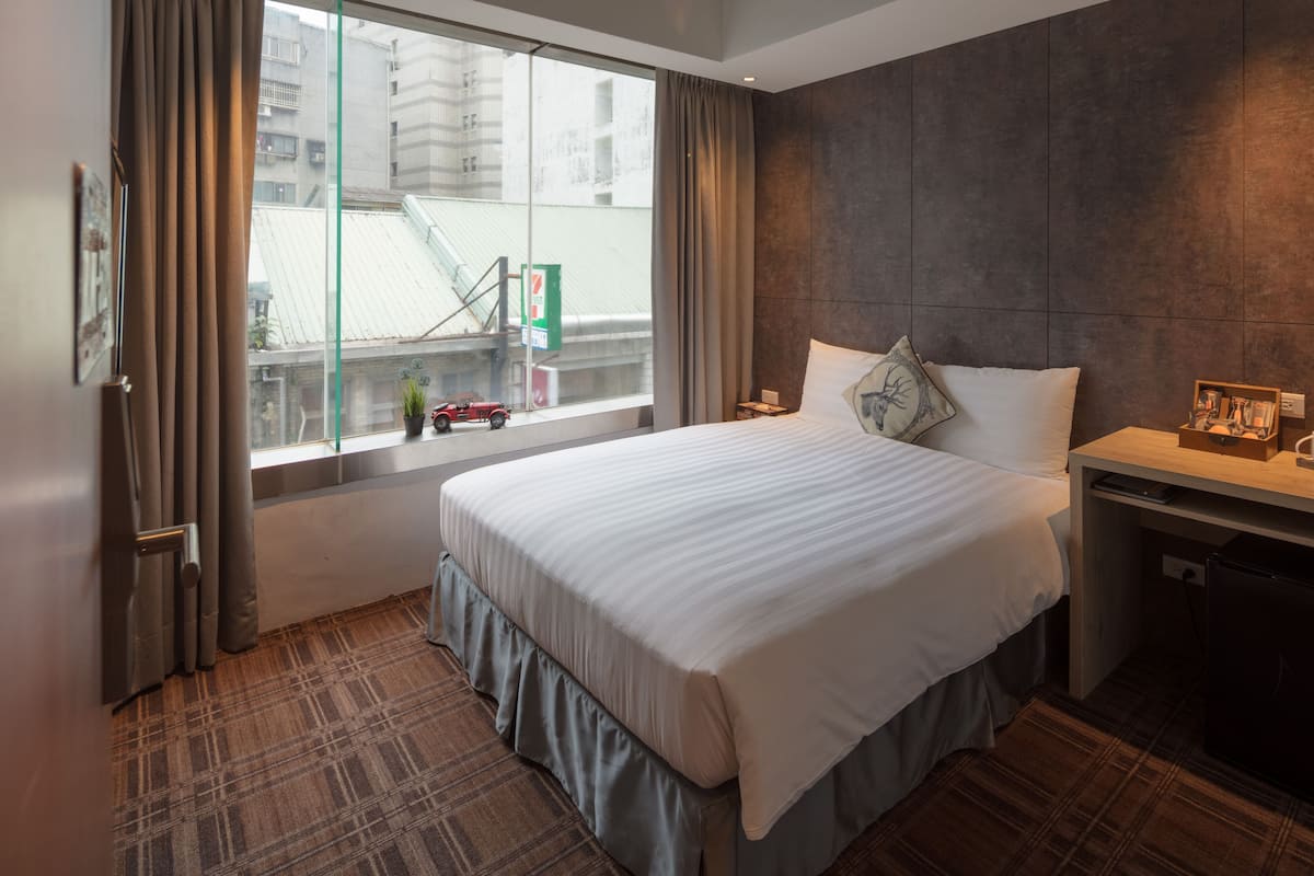 A hotel room with a large bed, a desk, a window with curtains, and a view of buildings outside.