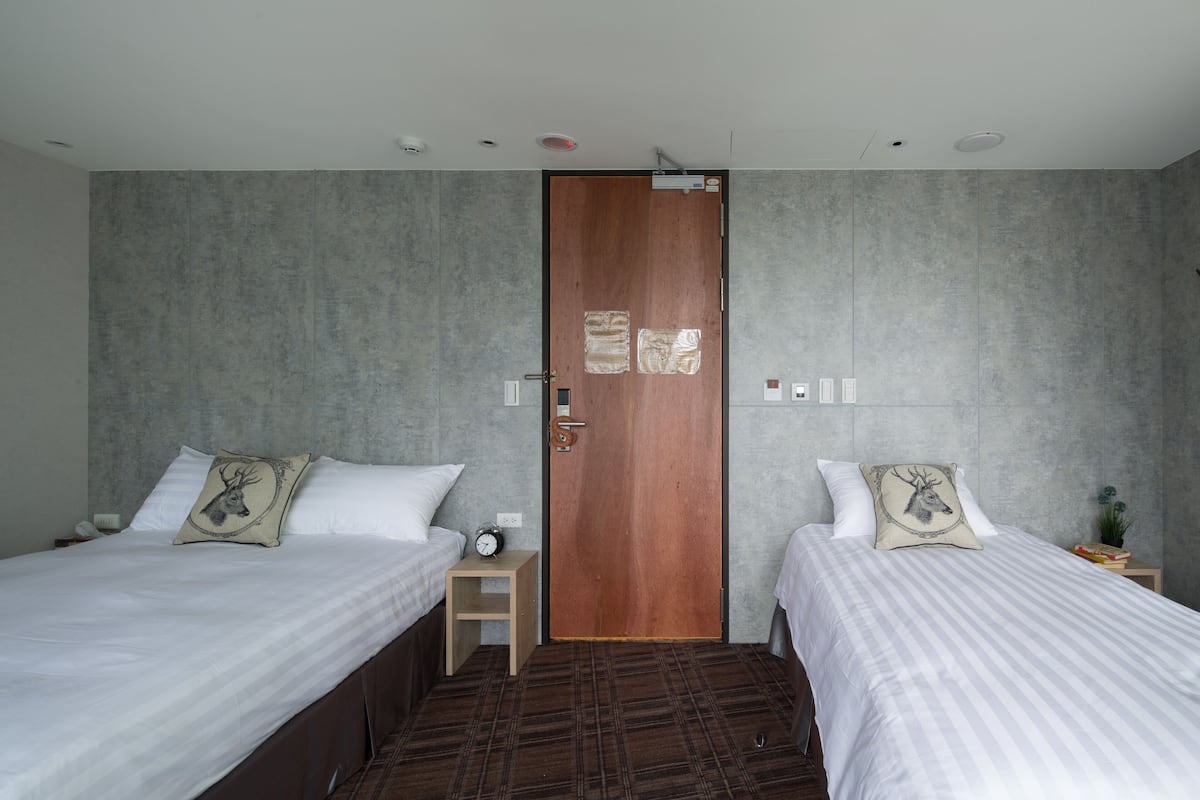 A hotel room with two beds, a wooden door, and a small bedside table with a clock.