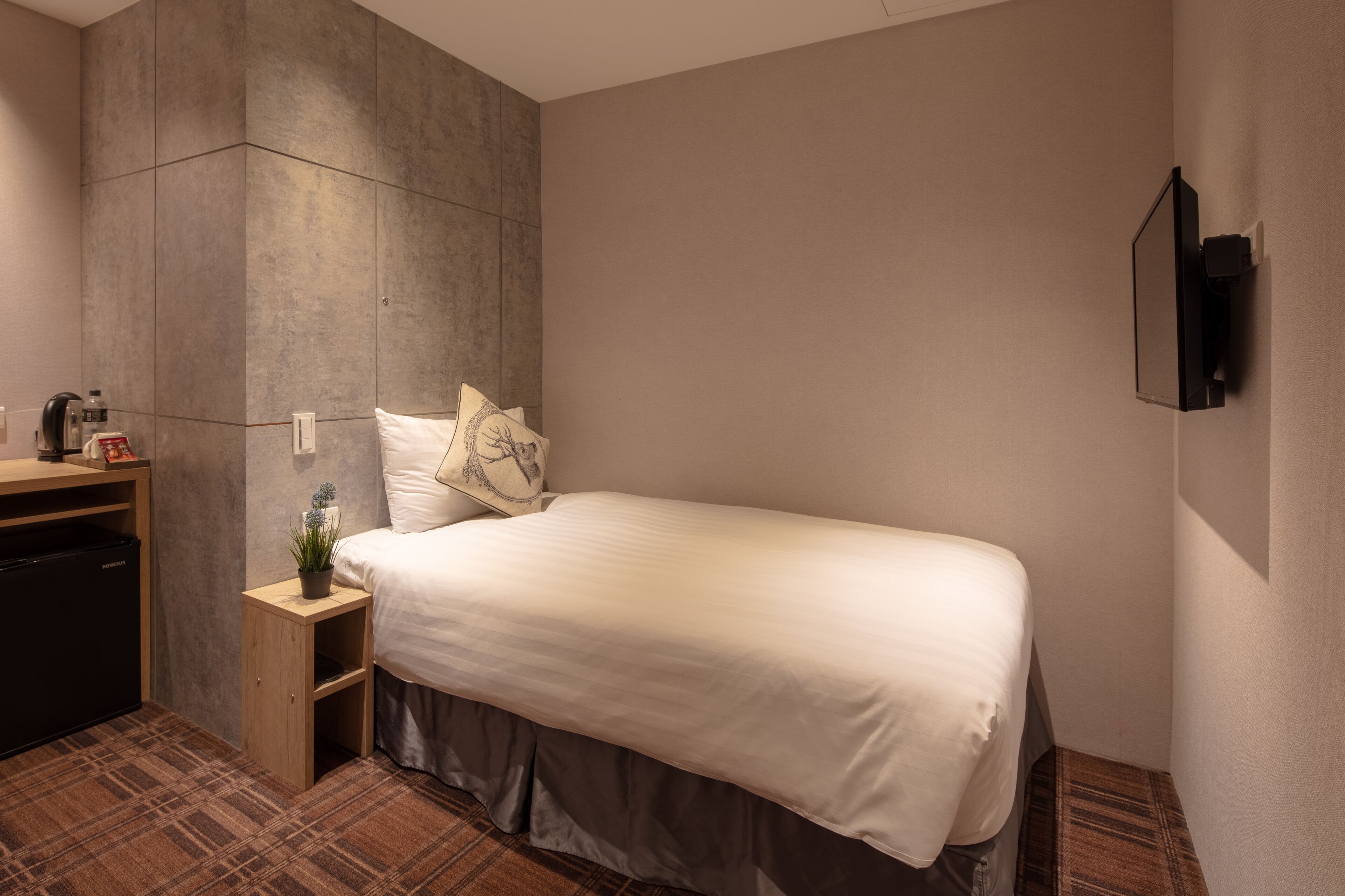A hotel room with a bed, a small bedside table with a plant, a wardrobe, and a built-in TV.