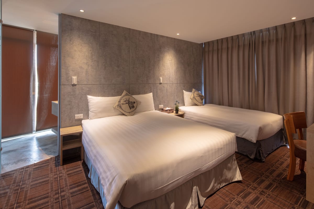 A hotel room with two beds, a wooden chair, a small table, and a large window with curtains.