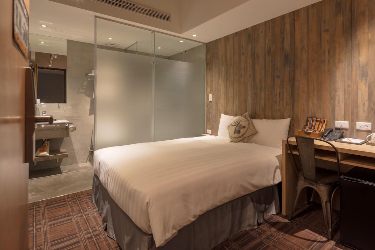 A modern hotel room with a large bed, a desk with a chair, a small refrigerator, a wooden accent wall, and a glass shower enclosure.