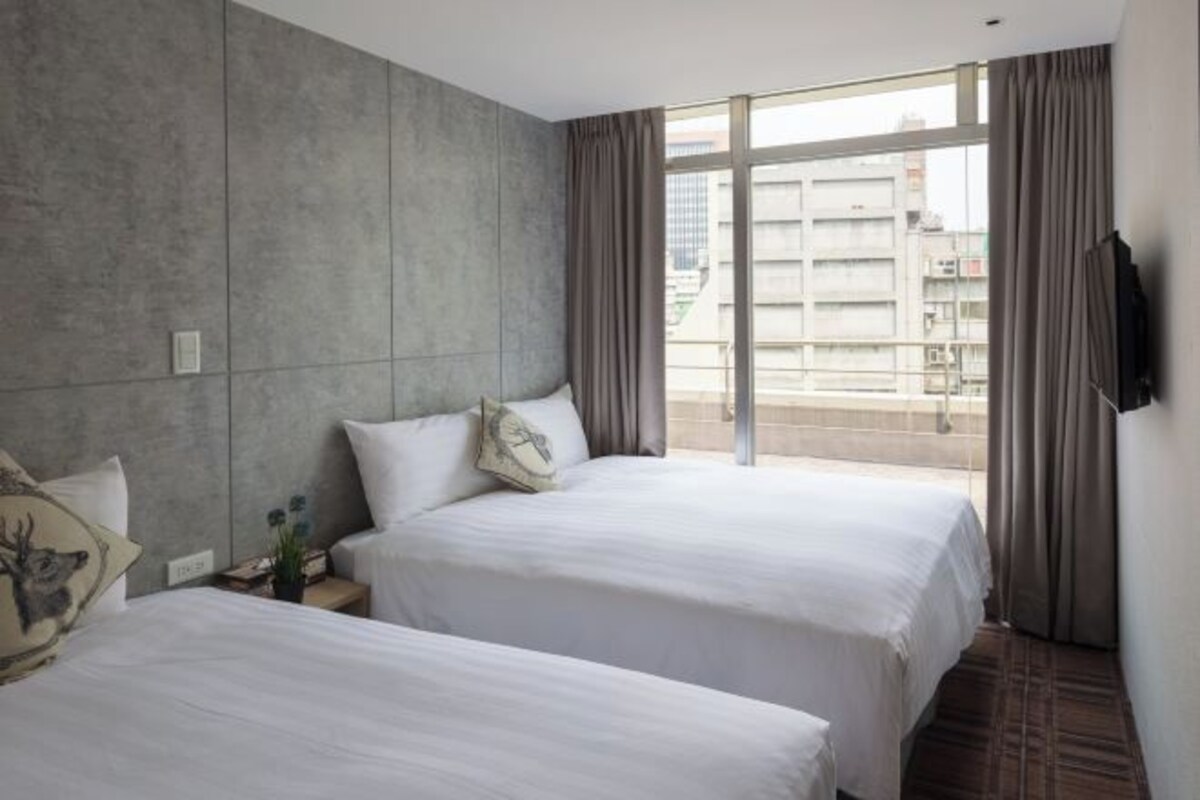 A hotel room with two beds, a large window with curtains, and a view of buildings outside.