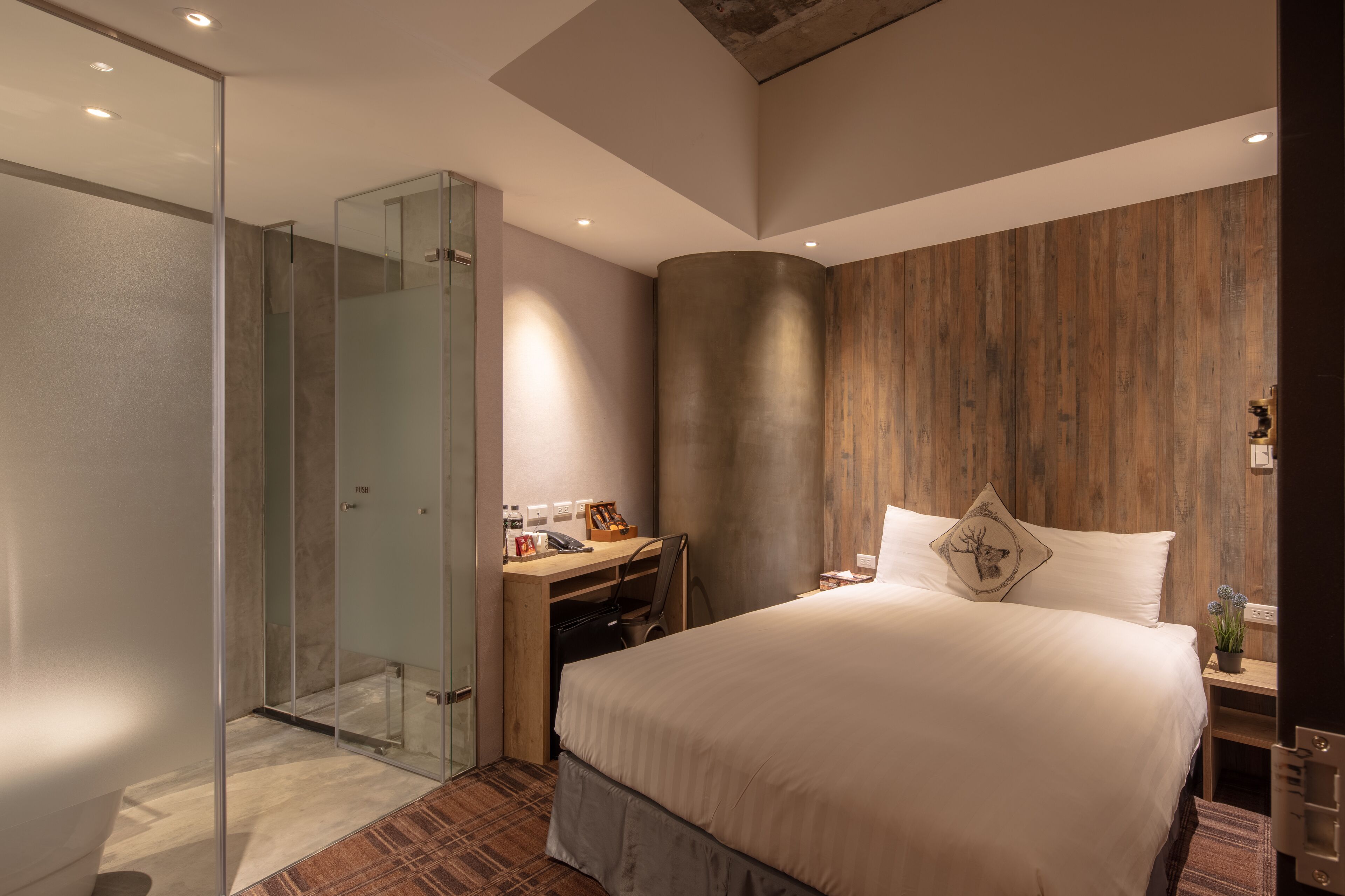 A modern hotel room with a large bed, a desk, a glass shower enclosure, and a wooden accent wall.