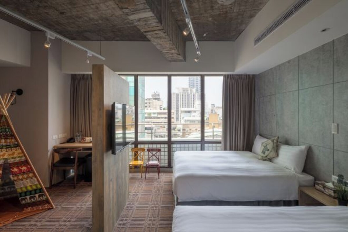 A hotel room with two beds, a desk, a chair, and a large window offering a city view.