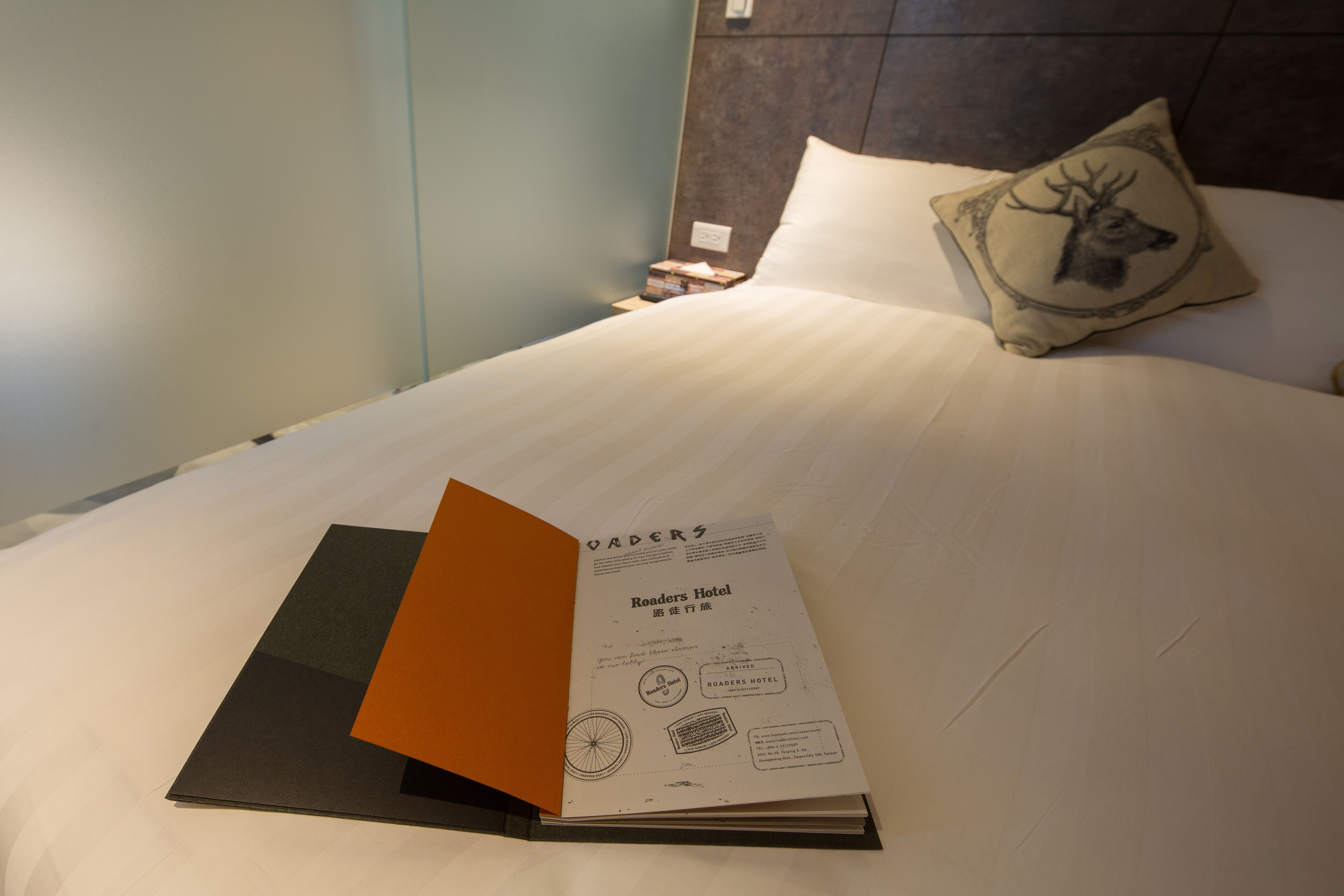 Cama bem arrumada com travesseiro de desenho de cabeça de veado, um livro intitulado "Roaders Hotel" sobre a cama e uma mesa de cabeceira com uma luminária.
