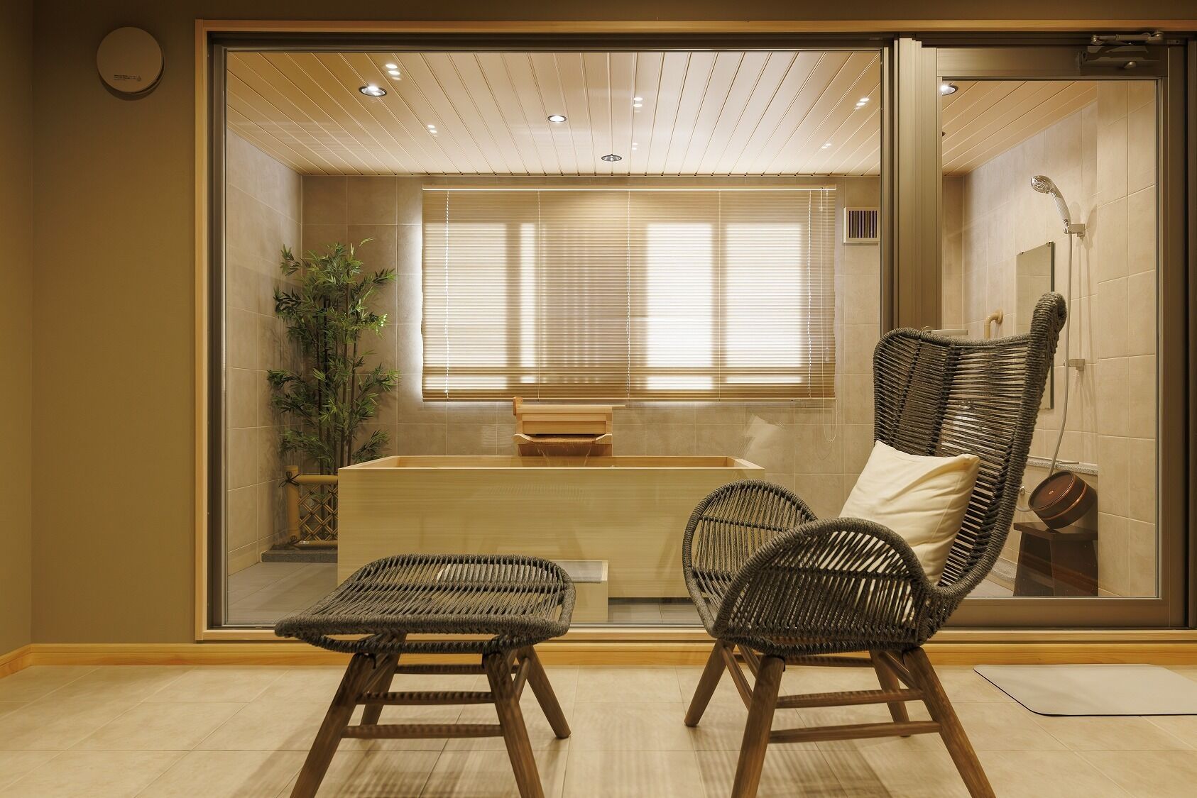 A room with a wooden bench, two wicker chairs, a wooden table, and a large window with blinds.