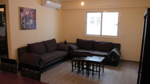 Una sala de estar con un sofá modular, una mesa de comedor y sillas. Se aprecia una ventana con vista a edificios.