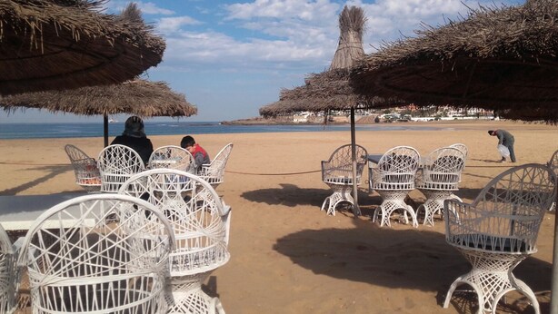 Un café frente al mar con sillas de mimbre blanco y sombrillas de paja.