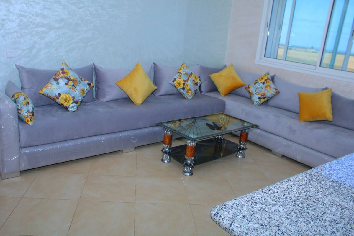 Sala de estar com sofá cinza, almofadas estampadas em amarelo e floral, mesa de centro de vidro e vista pela janela.