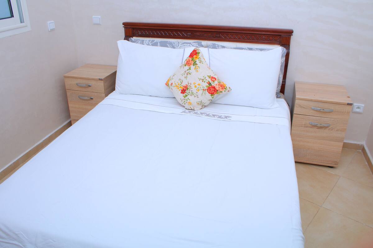 Cama bem arrumada com travesseiro floral, cabeceira de madeira e duas mesas de cabeceira com gavetas.