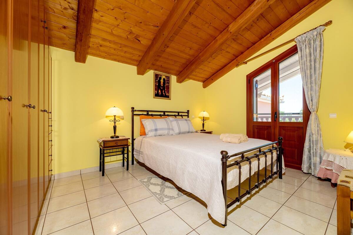 A bedroom with a wooden ceiling, a bed with two bedside tables, a window with curtains, and a door.