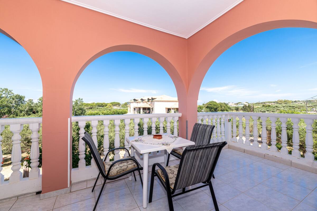 A balcony with a white table and chairs, a white railing, and a view of a building and greenery.