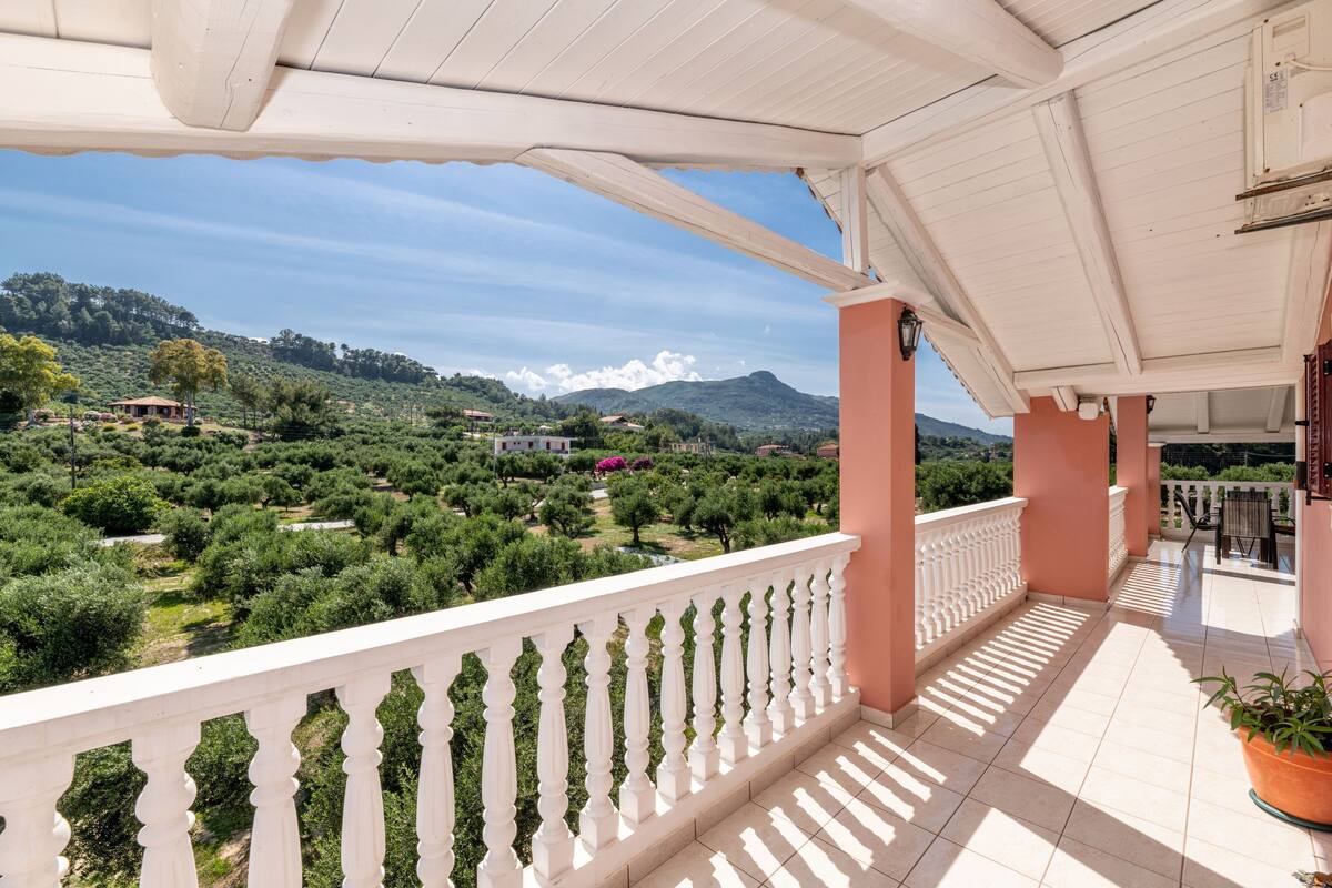 A balcony with a white balustrade overlooking a lush landscape with trees and distant mountains.