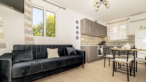 A modern living room with a dark sofa, a dining area with a table and chairs, and a kitchen with wooden cabinets and a window. - Arazzo Apartments (Zakynthos)