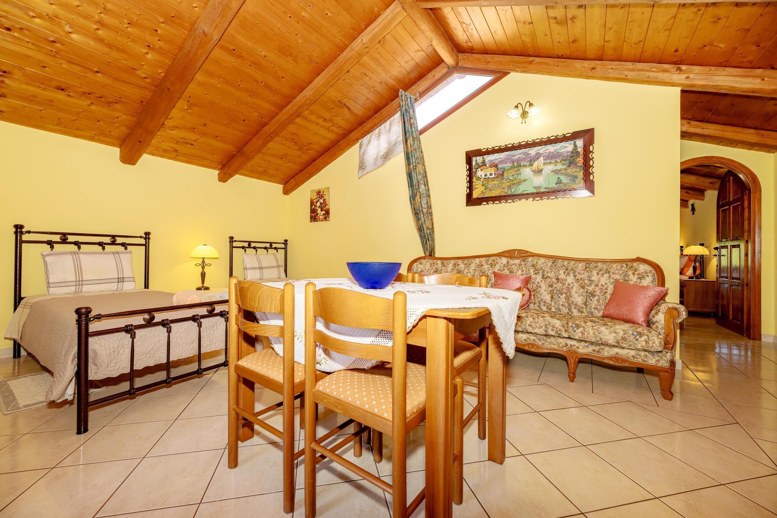 A room with a wooden ceiling, a bed, a dining table with chairs, a sofa, and a painting on the wall.