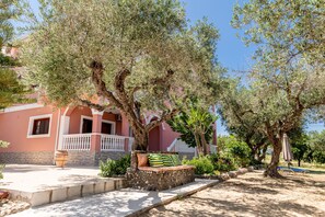 A stone bench with cushions, a potted plant, and a pergola under olive trees in a courtyard. - Arazzo Apartments (Zakynthos)