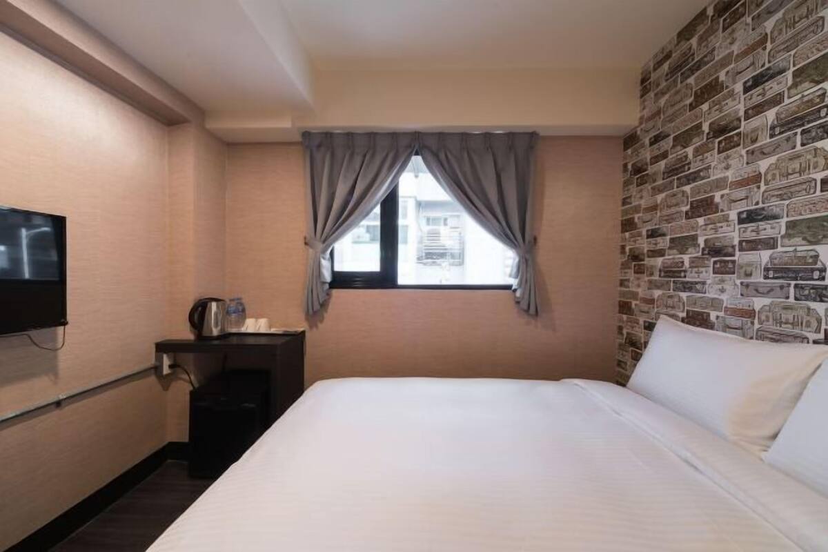 A hotel room with a bed, a TV, a small table with a kettle, and a window with curtains.