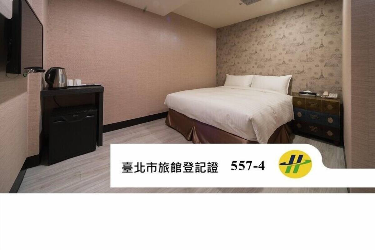 A hotel room with a bed, a small desk with a kettle, and a nightstand with a lamp.
