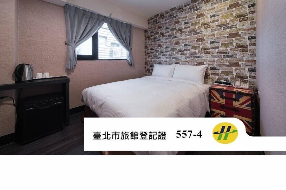 A hotel room with a brick-patterned wall, a bed with white linens, a nightstand with a suitcase, and a window with curtains.