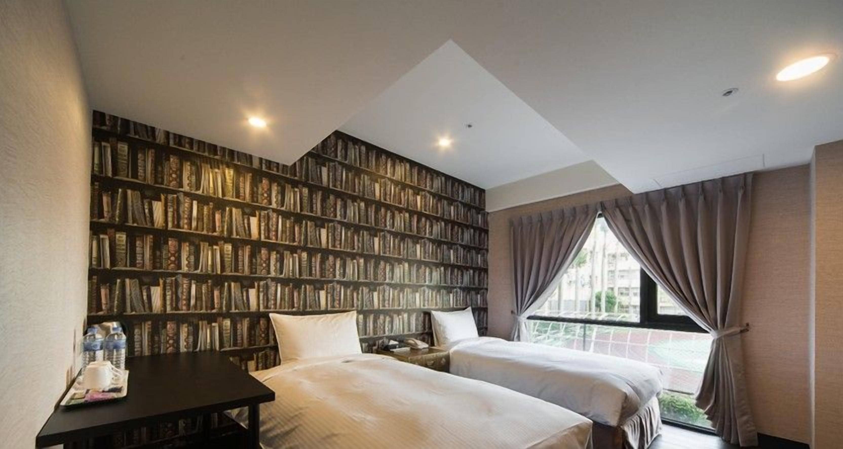 A hotel room with two beds, a bookshelf, a desk, and a window with curtains.