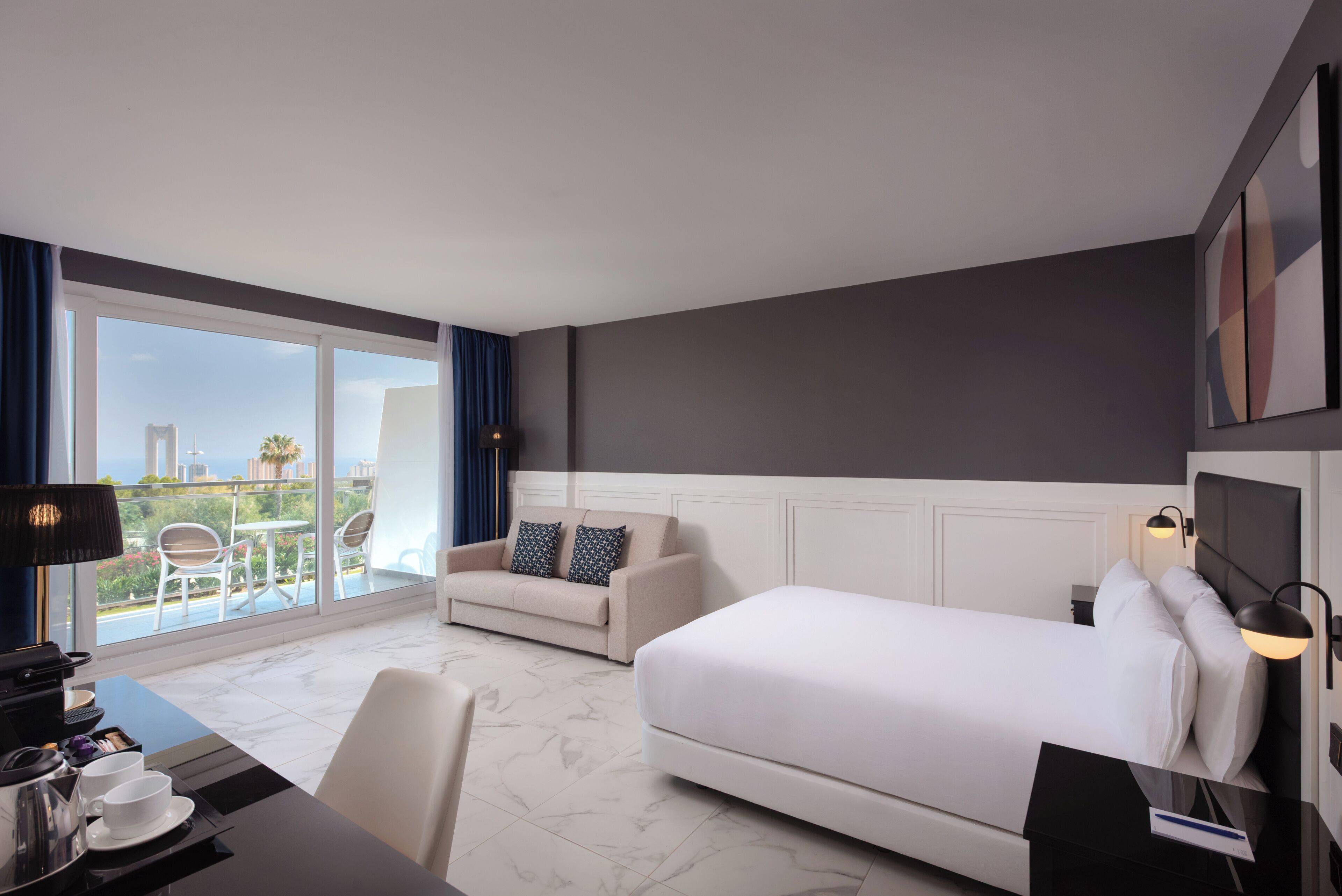 A hotel room with a bed, a sofa, a desk with a lamp, a coffee machine, and a view of the city.