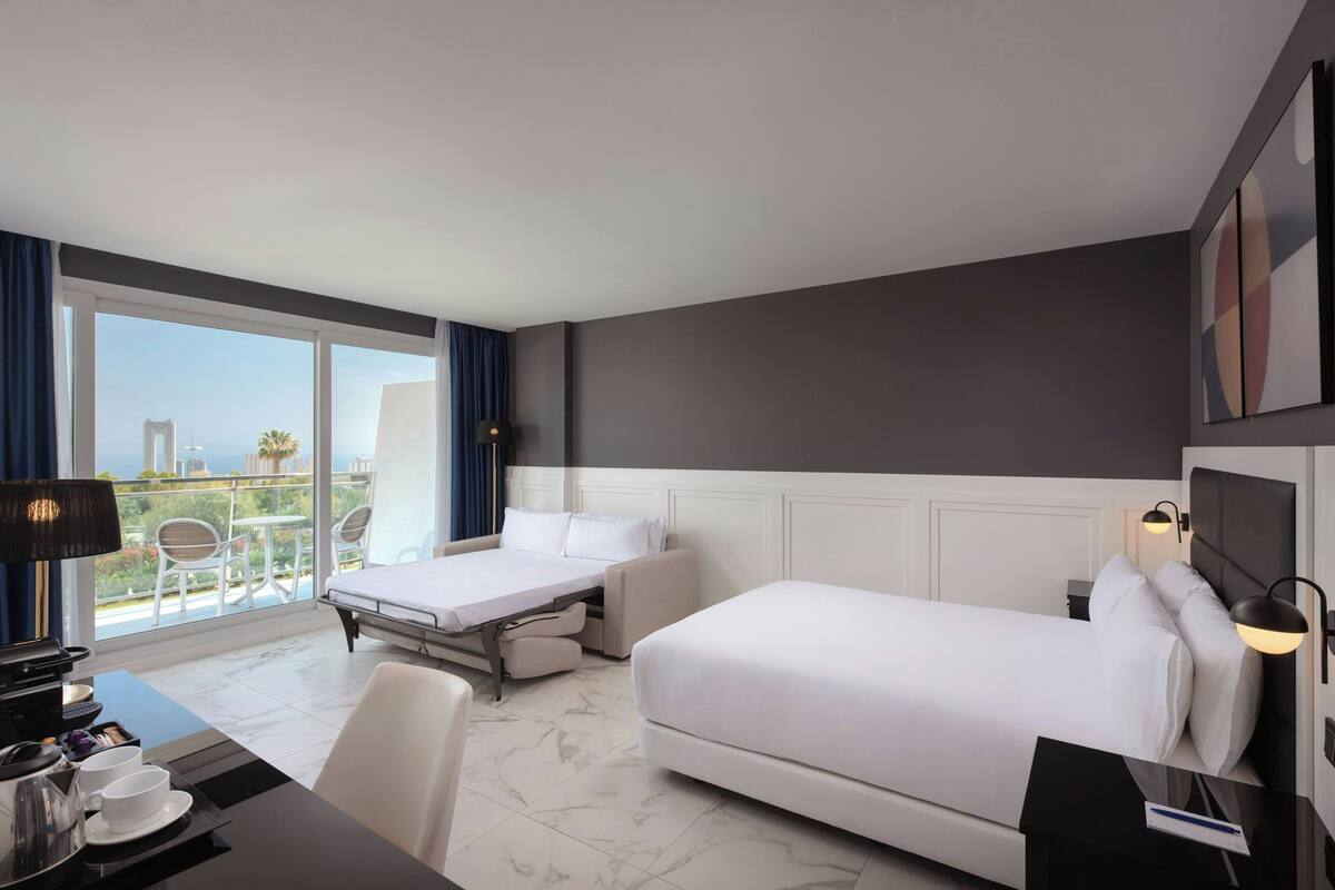 Habitación de hotel con cama, escritorio, silla, mesa con tazas y bandeja, y balcón con sillas y mesa.