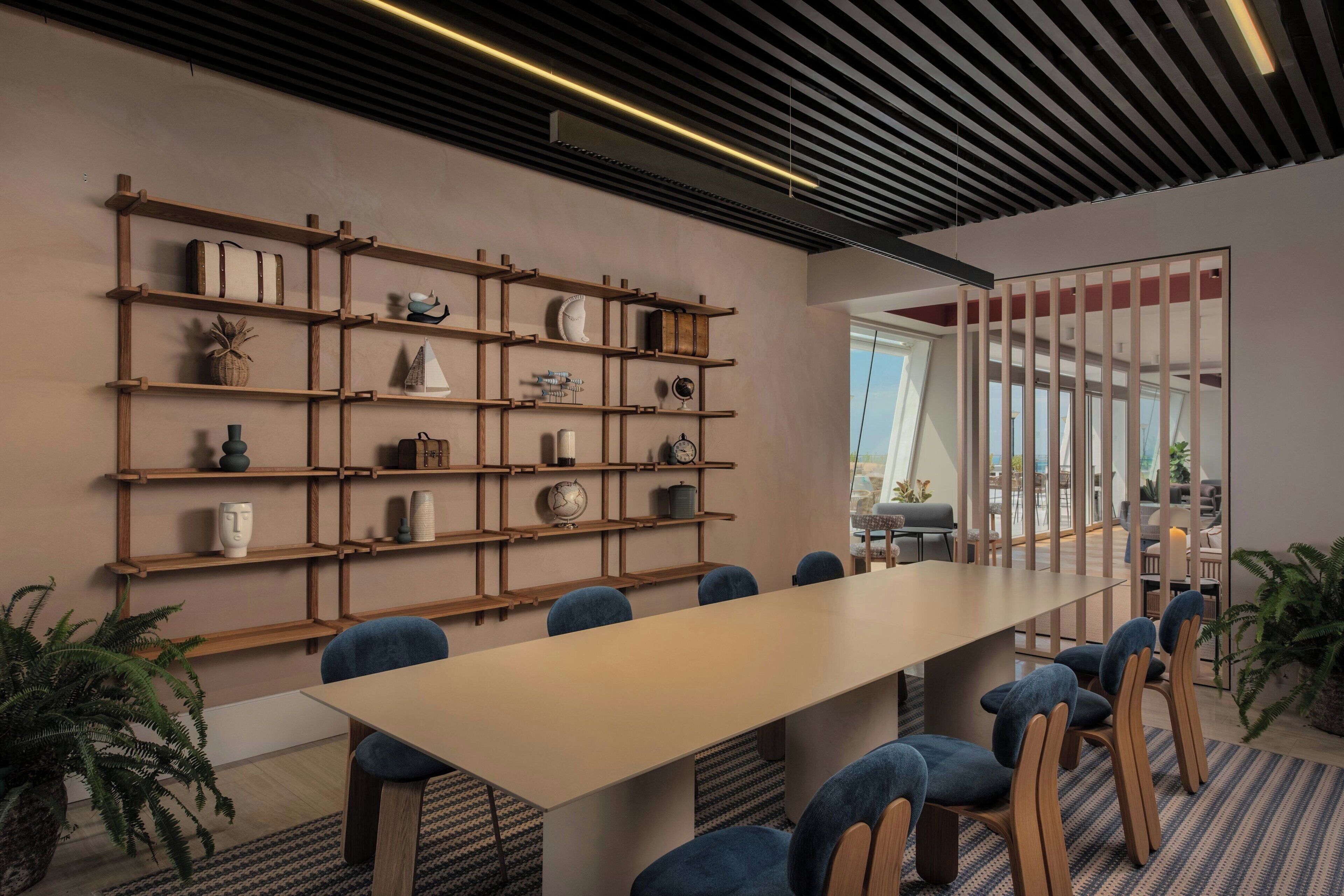 A modern meeting room with a long table, blue chairs, wooden shelves with decorative items, and a view through a glass partition.