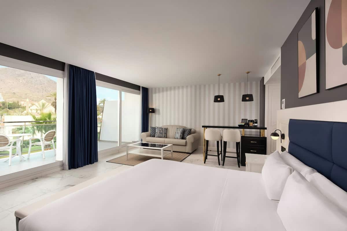 Una habitación de hotel moderna con una cama grande, un área de comedor y un balcón con vistas a las montañas.