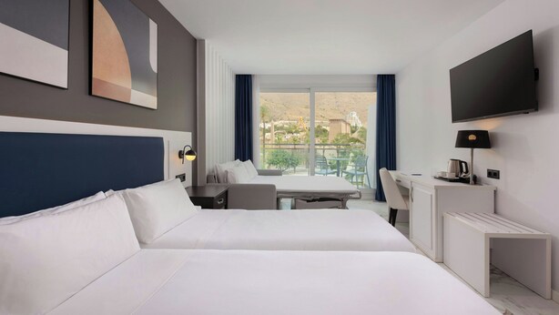Habitación de hotel con dos camas, un escritorio, una silla, un televisor y un balcón con vistas a la ciudad.