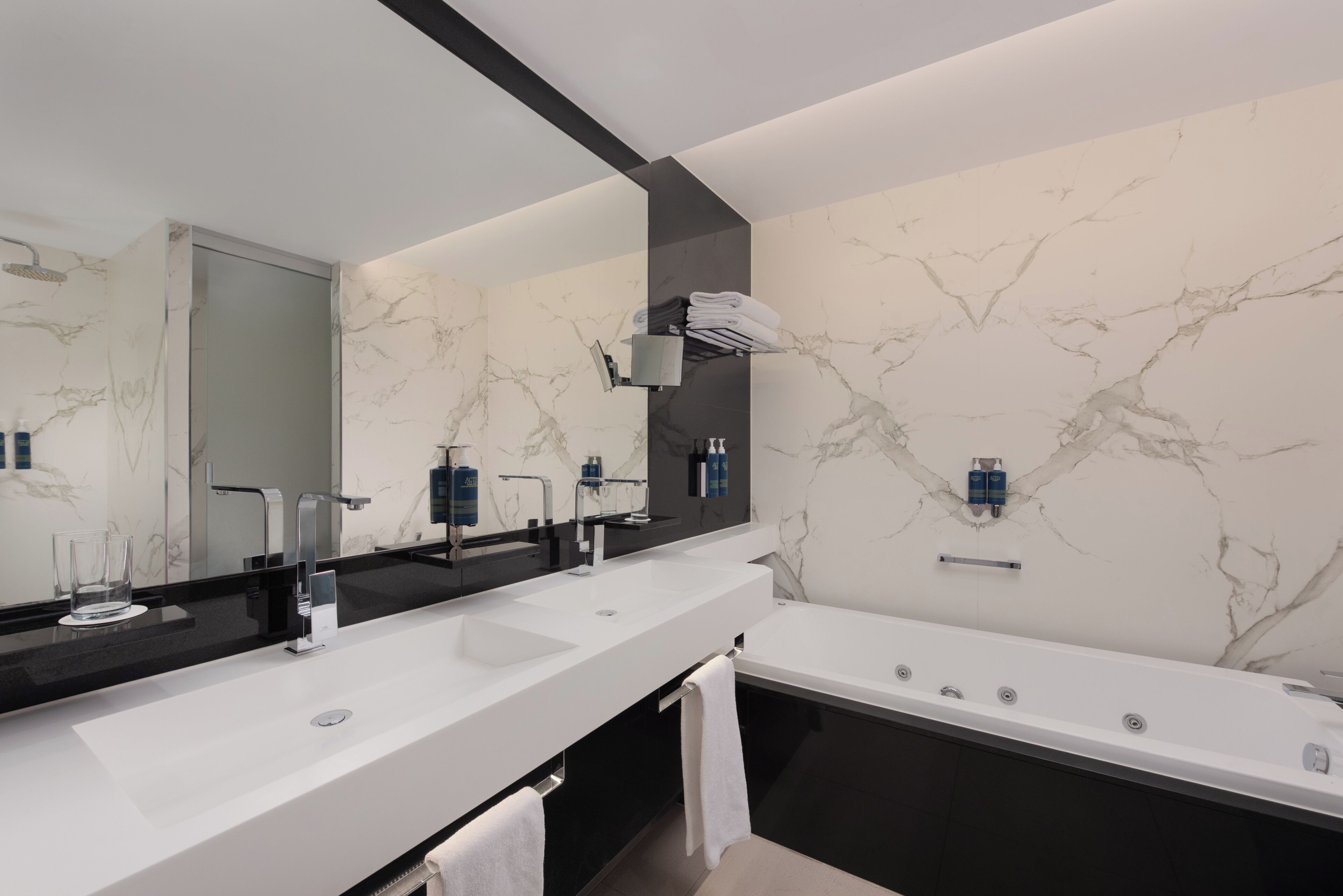 A modern bathroom with a large mirror, two sinks, a bathtub, and marble-patterned walls.