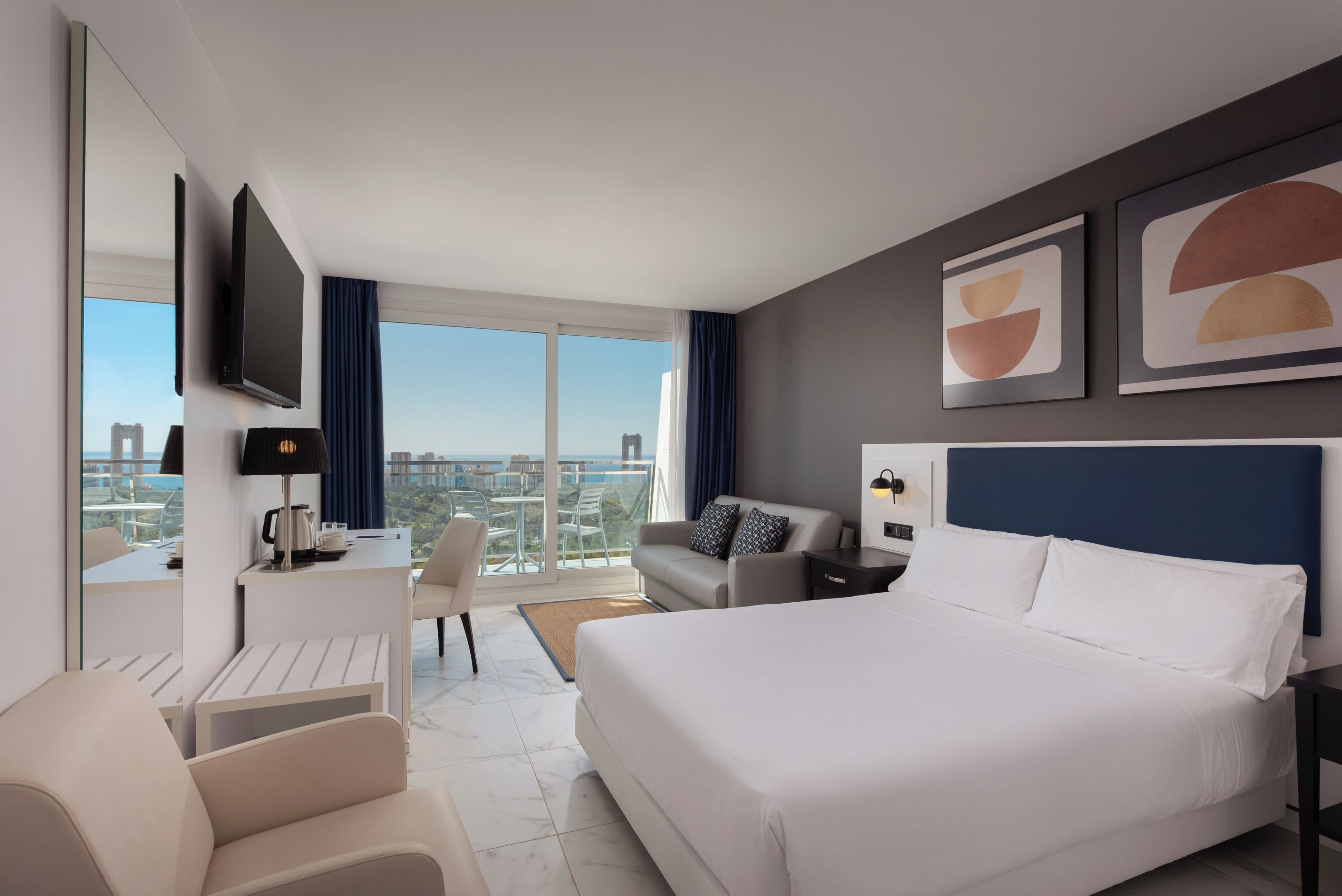 Ein modernes Hotelzimmer mit einem großen Bett, einem Schreibtisch mit Stuhl, einer Sitzecke und einem an der Wand befestigten Fernseher. Blick auf die Stadt.