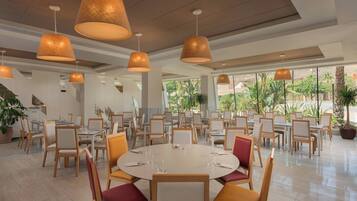Un vaste espace dédié à la salle à manger, avec des tables et des chaises rondes, de grandes lampes suspendues et une vue sur la verdure environnante.